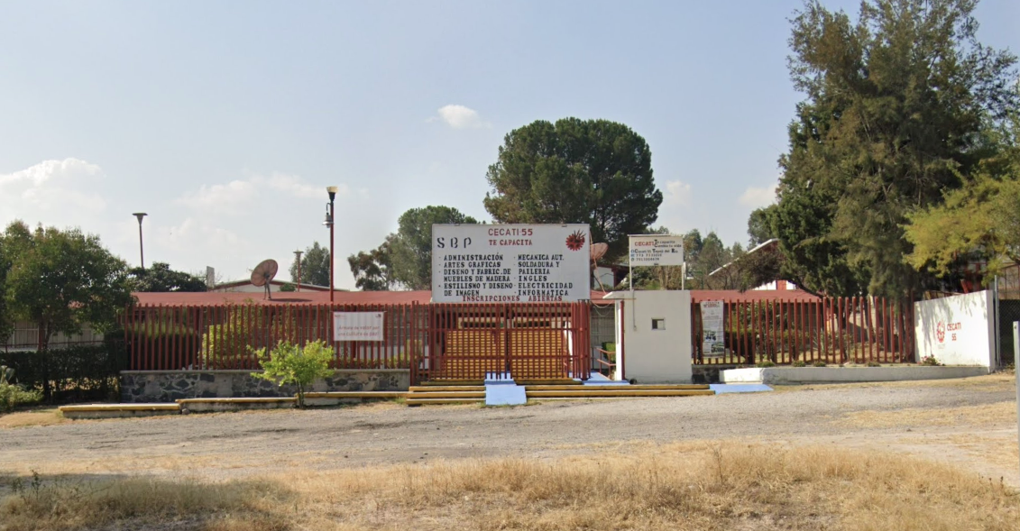 Fotografía en la que se muestra la entrada del plantel, conformada por una reja de color rojo. Sobre la reja hay una placa que contiene los datos del plantel, como el nombre y las carreras que se imparten. Frente a la reja hay unas escaleras y una rampa de acceso.